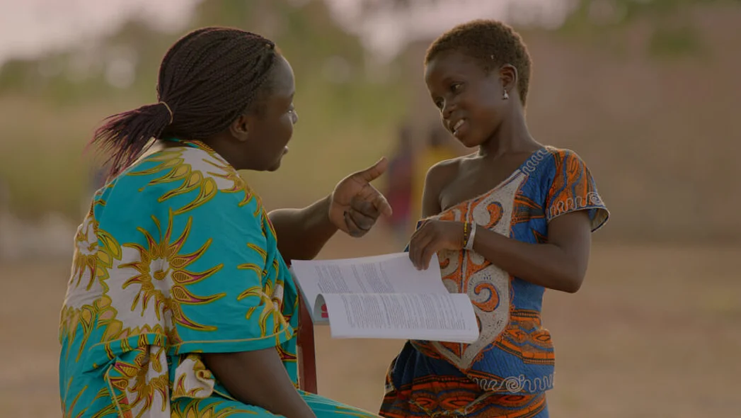Eine junge Frau unterh&auml;lt sich mit einem M&auml;dchen, das ihr ein Schulheft zeigt.