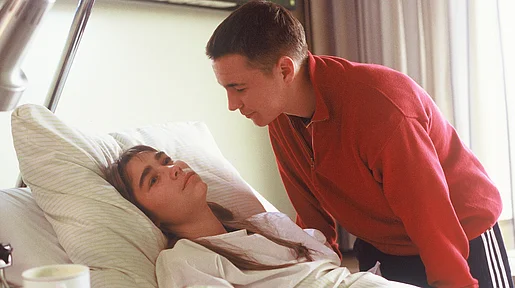 Szenenbild: ein junger Mann beugt sich über eine junge Frau, die in einem Krankenhausbett liegt.