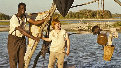 Szenenbild. Huck und Jim auf einem Floß in der Abendsonne