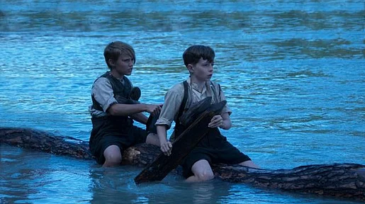 Szenenbild: Tom und Hacke sitzen auf einem Baumstamm und paddeln durchs Wasser, Abendstimmung