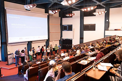 Blick von hinten in einen gro&szlig;en H&ouml;rsaal, auf dem Podium mehrere Menschen auf St&uuml;hlen