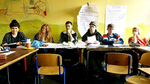 Szenenbild: Blick in Klassenraum, auf die hintere Reihe U-förmig aufgestellter Tische, die Wand ist gelb