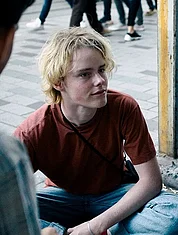 Standbild aus dem Film R&auml;uberh&auml;nde: die beiden Hauptfiguren sitzen auf dem Boden und unterhalten sich mit anderen, die angeschnitten im  Bildvorgergrund zu sehen sind.
