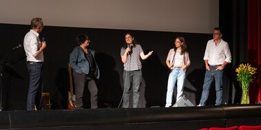 Blick auf die Bühne eines Kinos, fünf Menschen stehen dort, eine Frau mit Mikrofon in der Hand