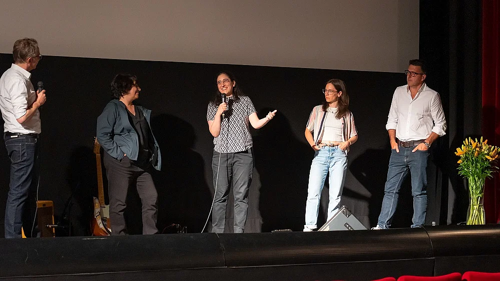 Blick auf die B&uuml;hne eines Kinos, f&uuml;nf Menschen stehen dort, eine Frau mit Mikrofon in der Hand