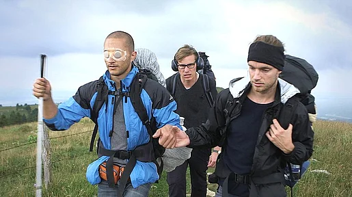 Szenenbild: die drei Freunde mit Wanderstab, einer mit zugeklebten Augen, einer mit Kopfhörern, auf einer Wiese in Outdoorklamotten