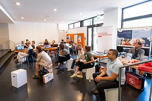 Eine Gruppe von Menschen, auf Pappschemeln verteilt in einem gro&szlig;en Raum