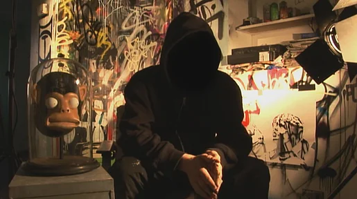 Szenenbild: Mann mit Kapuze auf, so dass sein Gesicht nicht zu sehen ist, vor Graffitti Wand