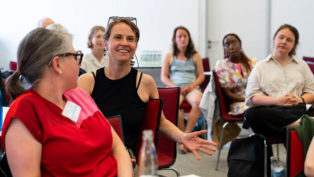 eine l&auml;chelnde Frau in der linken Bildmitte, mehrere Personen drumherum, im Vordergrund dreht sich eine Frau zu der l&auml;chelnden Frau um