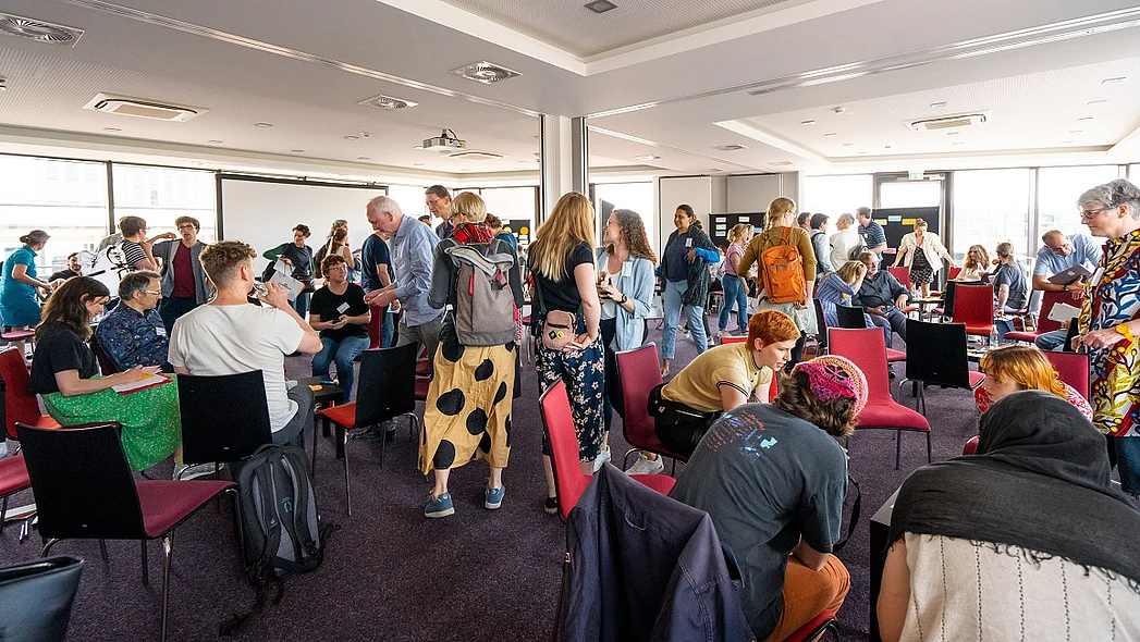 Sehr viele Menschen stehend und sitzend in einem gro&szlig;en Raum verteilt, zum Teil im Gespr&auml;ch