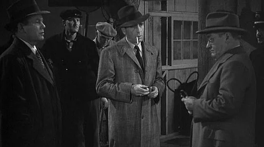 Szenenbild: Eine Gruppe von Männern, unter ihnen Humpfrey Bogart als Philipp Marlow in Trenchcoat und mit Hut