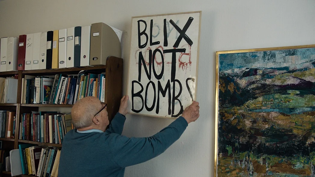 Ein &auml;lterer Mann h&auml;ngt ein Bild zwischen einem B&uuml;cherregal und einem &Ouml;lgem&auml;lde ab oder auf, auf dem Blix not Bombs zu lesen ist.