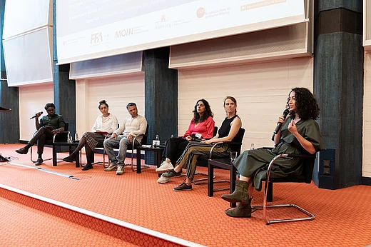 Sechs Menschen auf einer B&uuml;hne vor einer Leinwand, eine Frau spricht in ein Mikrofon