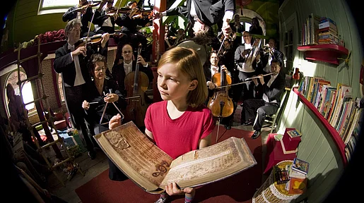 Lili hält ein Buch aufgeschlagen in ihren Händen. Im Hintergrund ist ein Orchester. 