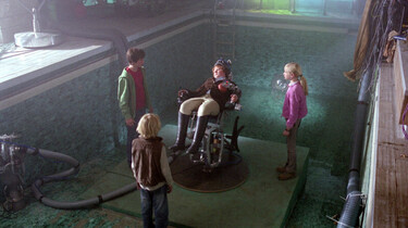 Szenenbild: Tim, Karl und Gaby stehen um einen experimentellen Stuhl auf dem Klößchen liegt