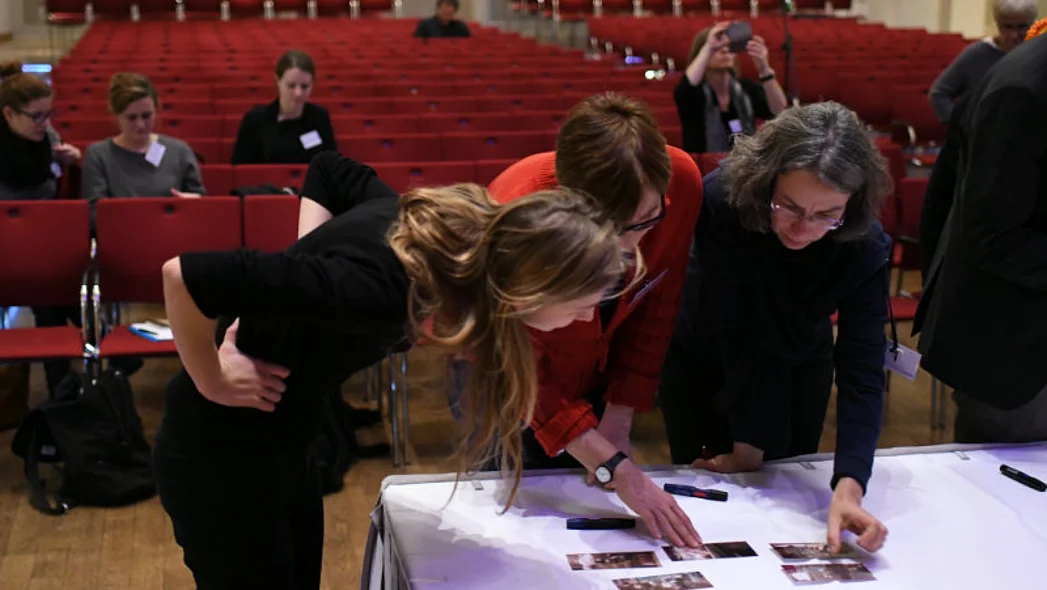 Drei Frauen beugen sich &uuml;ber einen Tisch, auf dem Bilder liegen, im Hintergrund Sitzreihen