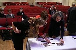 Drei Frauen beugen sich &uuml;ber einen Tisch, auf dem Bilder liegen, im Hintergrund Sitzreihen