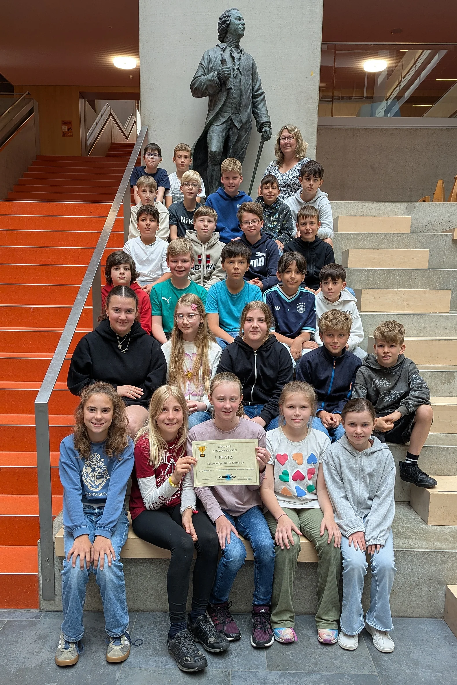 Alle Sch&uuml;ler*innen sitzen mit ihrer Klassenlehrerin auf einer gro&szlig;en Treppe und pr&auml;sentieren ihre Urkunde.
