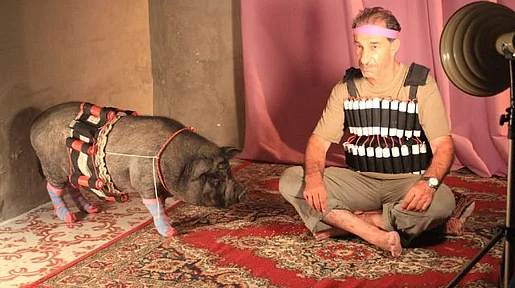 Szenenbild: Mann und Schwein mit Sprengstoffgürtel auf einem Teppich im Scheinwerferlicht
