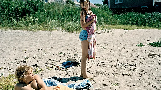 Szenenbild: am Strand: Ein Mädchen im  Badezug schaut auf einen am Boden liegenden Jungen