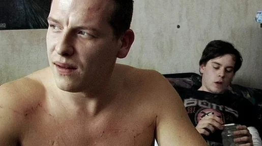 Szenenbild: Mann mit nacktem Oberkörper im Bildvordergrund, im Hintergrund junger Mann  auf Sofa