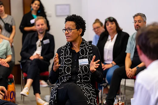 Rednerin Mo Asumang auf einem Stuhl sitzend, im Hintergrund Teilnehmende in Unsch&auml;rfe