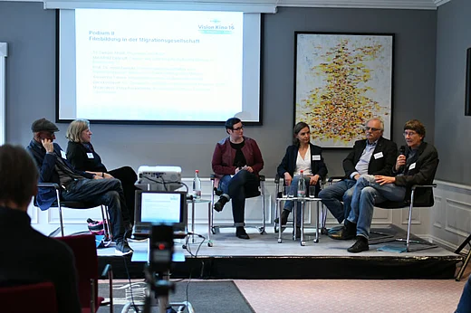 Blick auf ein Podest, auf dem die Podiumsteilnehmenden sitzen, im Hintergrund Leinwand