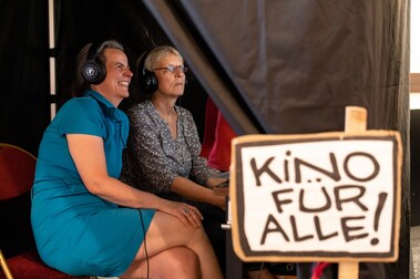 In der linken Bildhälfte zwei lachende Frauen mit Kopfhörern, in der rechten Bildhälfte ein Plakat, auf dem "Kino für alle" steht