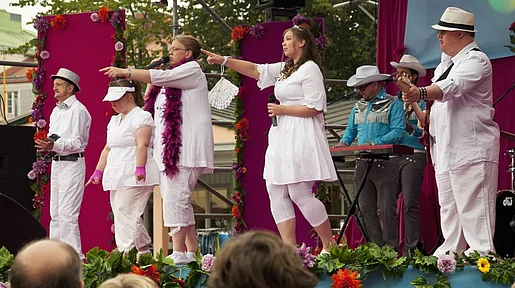Szenenbild: Die Theatergruppe auf einer Freiluftbühne, weiß gekleidet vor pinkem Hintergrund