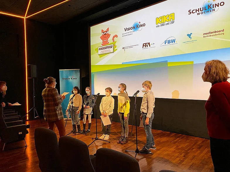 f&uuml;nf Kinder hinter Mikrofonen vor einer Kinoleinwand, schr&auml;g von vorn aufgenommen