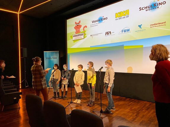 fünf Kinder hinter Mikrofonen vor einer Kinoleinwand, schräg von vorn aufgenommen