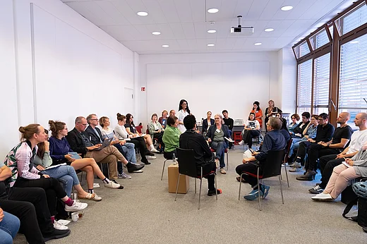 Vier Menschen in einem kleinen Stuhlkreis, drumherum viel mehr Menschen in einem gro&szlig;en Stuhlkreis