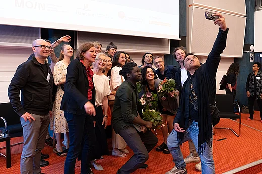 Eine gr&ouml;&szlig;ere Gruppe von Menschen auf einer B&uuml;hne, ein Mann, Hamze Bytyci, macht ein Selfie von allen