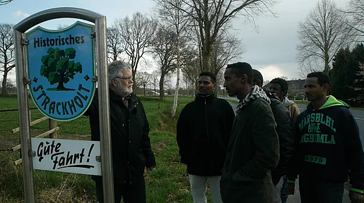 Szenenbild: Die Gruppe von Flüchtlingen steht mit Begleiter aus dem Ort an einem Ortsschild