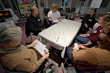 Um einen Tisch herum sind mehrere Personen im Gespr&auml;ch