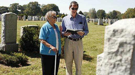 Szenenbild: Eine alte Frau mit Stock steht auf einem Friedhof neben einem Mann, der ein Buch in der Hand hält