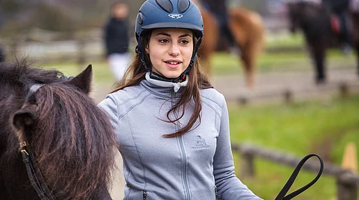 Szenenbild: Mädchen mit Reiterhelm führt ein braunes Pferd, dessen Kopf zu sehen ist