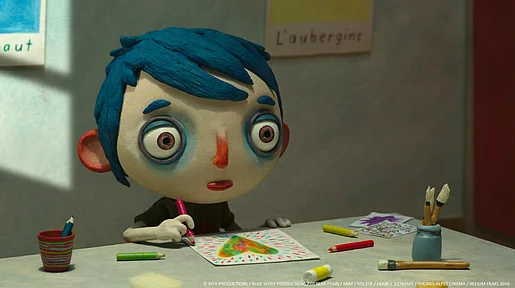 Szenenbild aus dem Puppentrickfilm: Zucchini sitzt an einem Tisch und malt ein Bild. Die Figur hat einen übergroßen Kopf, riesigen Augen und blaue Haare