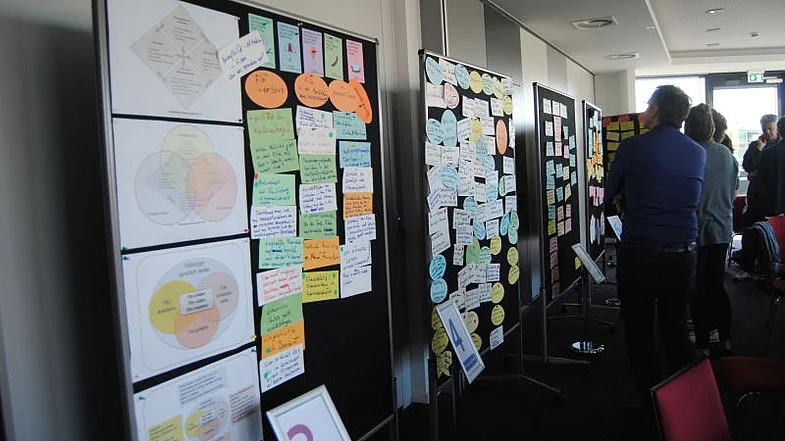 Pinw&auml;nde mit bunten Zetteln best&uuml;ckt erstrecken sich vom linken Bildrand bis zur Bildmitte, in der rechten Bildh&auml;lfte im Hintergrund mehrere Personen