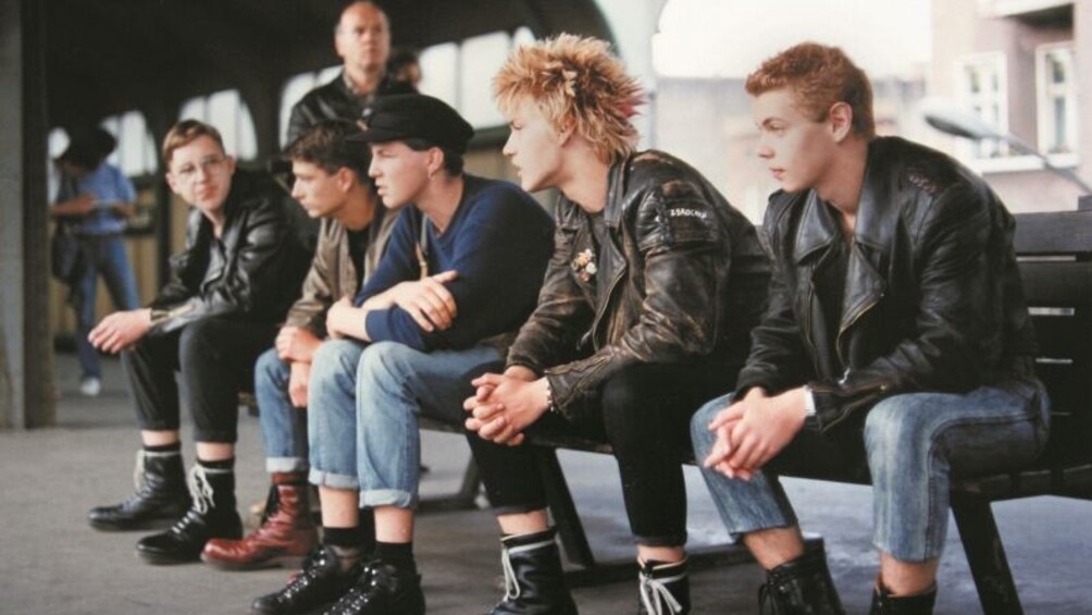 Fünf Musiker sitzen nebeneinander auf einer Band an einem Bahngleis.