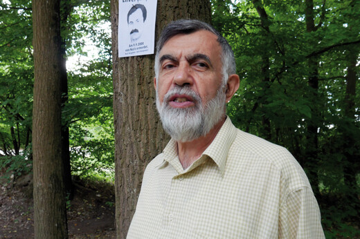 Ein älterer Mann steht vor einem Baum, an dem ein Plakat angebracht ist