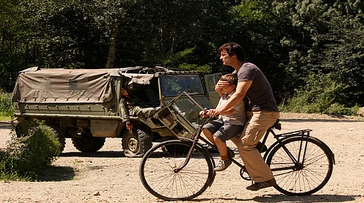 Szenenbild: Ein Mann und ein Kind auf einem Fahrrad, der Mann hält dem Kind die Augen zu, denn im Hintergrund ist ein Toter in einem Militär-Jeep zu sehen