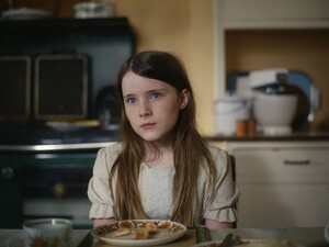Ein Mädchen sitzt in einer Küche am Tisch, vor ihr steht ein Teller. Sie blickt geradeaus. 