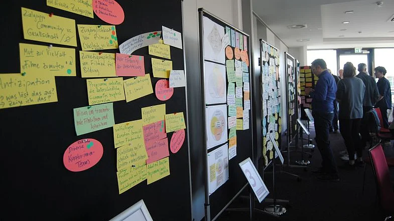 Pinw&auml;nde mit bunten Zetteln best&uuml;ckt erstrecken sich vom linken Bildrand bis zur Bildmitte, in der rechten Bildh&auml;lfte im Hintergrund mehrere Personen