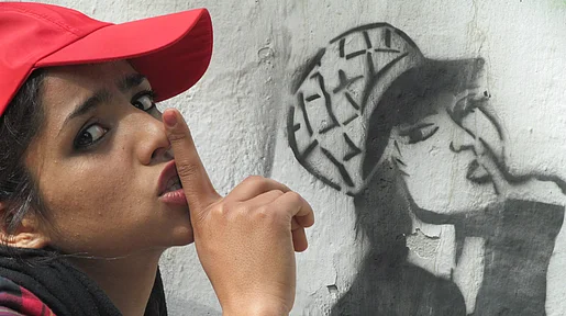 Szenenbild: Soniat mit Basecap und Finger über den Lippen vor einem Graffitti, das sie oder eine sehr ähnliche Person darstellt