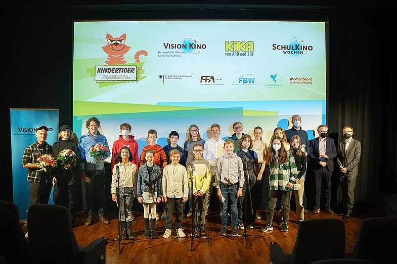 Gruppenbild aller Nominierten und Preistr&auml;ger:innen vor einer Kinoleinwand, auf der Logos zu sehen sind