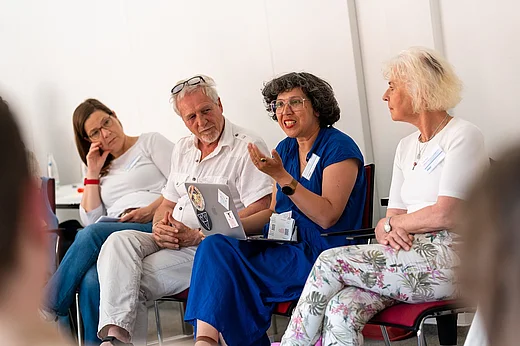 Vier Menschen sitzen auf St&uuml;hlen, eine Frau im blauen Kleid spricht in ein Mikrofon, die anderen sind ihr zugewandt