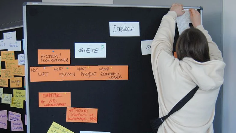 Eine Person heftet einen Zettel an eine Pinwand