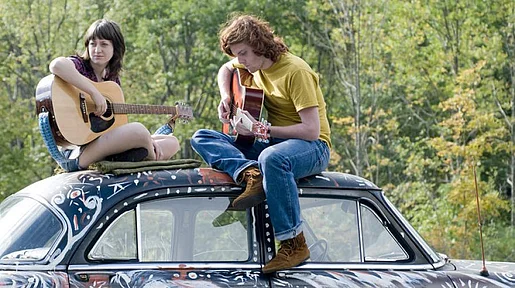 Szenenbild: Zwei junge Leute sitzen mit ihren Gitarren auf einem Autodach