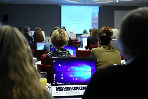 Blick auf Sitzreihen, man aufgeklappte Laptops, Menschen, im Hintergrund Leinwand